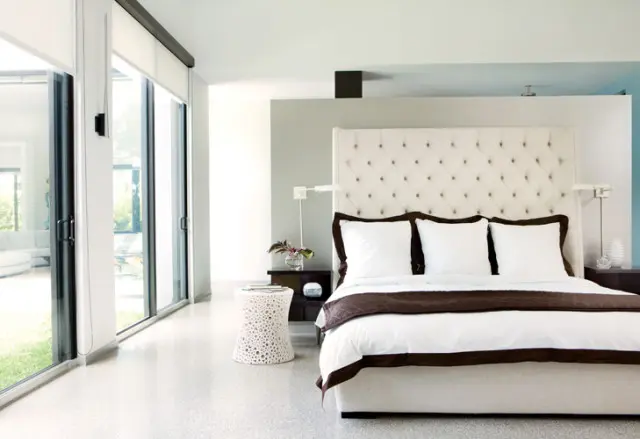 Modern bedroom with a tufted white headboard, white bedding with brown trim, and floor-to-ceiling windows overlooking a lawn.