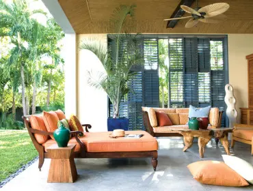 Covered patio with orange upholstered chaise lounge and sofa, natural wood coffee table, and large potted palm. Dark blue shutters are visible in the background.