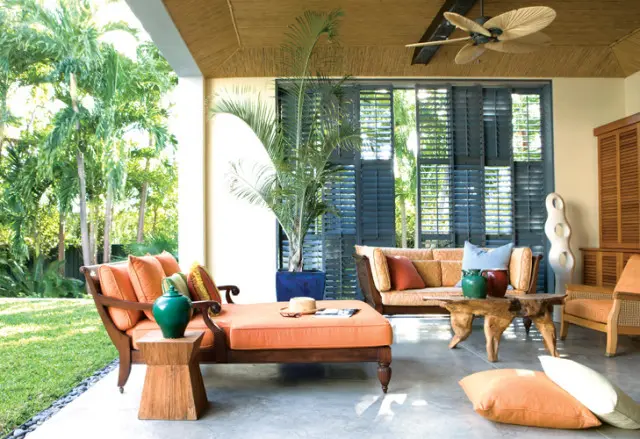 Covered patio with orange upholstered chaise lounge and sofa, natural wood coffee table, and large potted palm. Dark blue shutters are visible in the background.