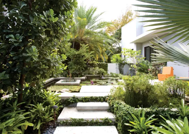 Modern concrete steps ascend through a lush garden with a water feature, palm trees, and a white modern house with an orange chair visible.