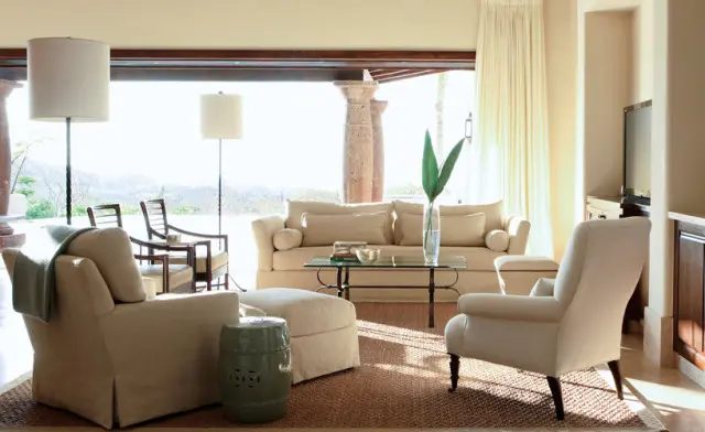 Living room with neutral-toned slipcovered sofas and armchairs, a glass-top coffee table, and two floor lamps. Large windows open to an outdoor patio with stone columns.