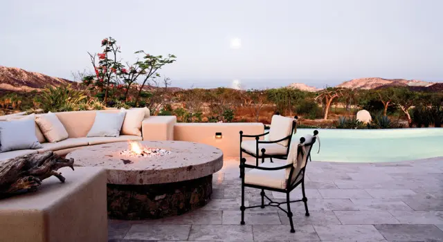 Terrace with built-in sectional sofa and stone fire pit overlooking a pool and arid landscape with mountains in the distance.