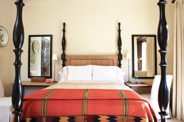 Bedroom with a dark wood four-poster bed, woven headboard, white bedding with orange trim, and a vibrant orange and patterned throw blanket.