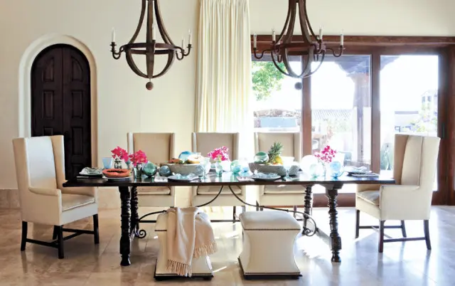 Dining room with a dark wood table set with fruit and flowers, flanked by upholstered chairs and two wooden chandeliers overhead.