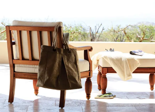 Wooden armchair with beige cushions and a canvas tote bag hanging from its arm, next to a bench with a folded towel, sunglasses, and a magazine.