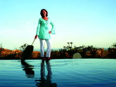 Woman in a light blue tunic and white pants stands at the edge of an infinity pool, holding a large green leaf.