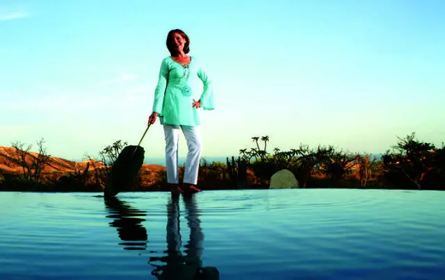 Woman in a light blue tunic and white pants stands at the edge of an infinity pool, holding a large green leaf.