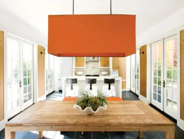 Dining area with a large wooden table, orange pendant light, and kitchen island with bar stools. White French doors line the walls.