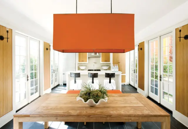 Dining area with a large wooden table, orange pendant light, and kitchen island with bar stools. White French doors line the walls.