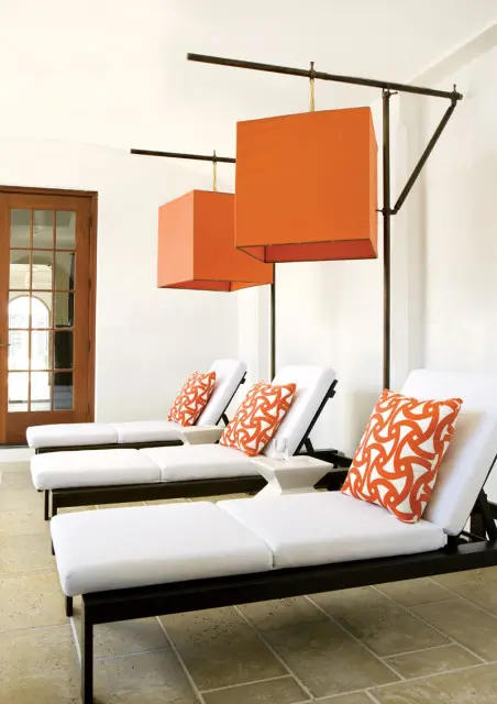 White chaise lounges with orange patterned pillows and large orange square pendant lights hanging above on a tiled patio with a wooden door.