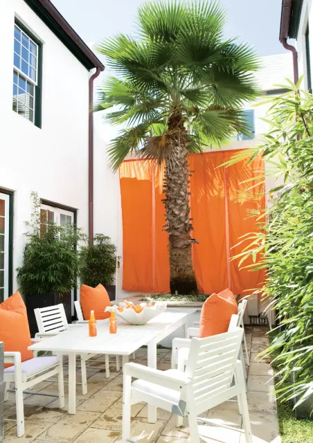 White patio furniture with orange cushions surrounds a white dining table on a stone patio, with a palm tree and orange drapery in the background.