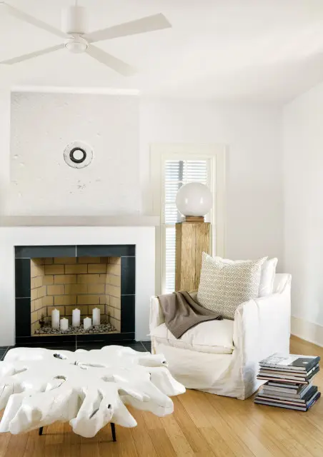 Living room with white slipcovered armchair, stacked books, white root coffee table, and fireplace with white candles.