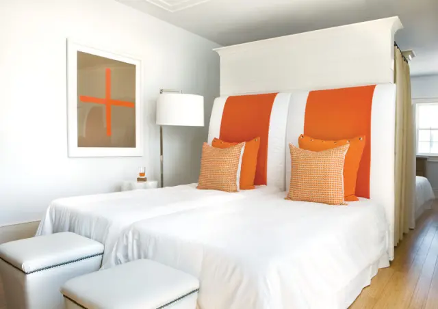 Bedroom with two twin beds covered in white linens, featuring orange and white striped headboards, orange accent pillows, and white ottomans at the foot of the beds.