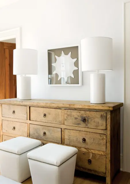 Bedroom with a rustic wooden dresser topped with two white cylindrical lamps and a framed starburst art piece. Two white upholstered ottomans sit in front of the dresser.