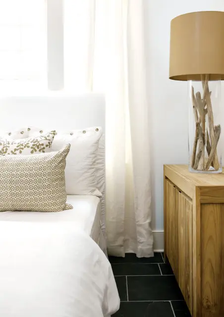 White upholstered headboard with white bedding and patterned pillows on a bed next to a wooden nightstand with a glass vase filled with driftwood and a tan lampshade.