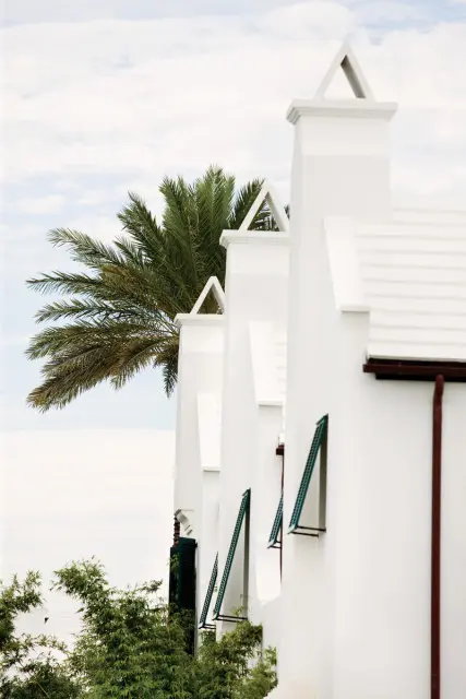 Row of white stucco buildings with triangular roof peaks and green shutters, featuring a palm tree and lush greenery against a cloudy sky.