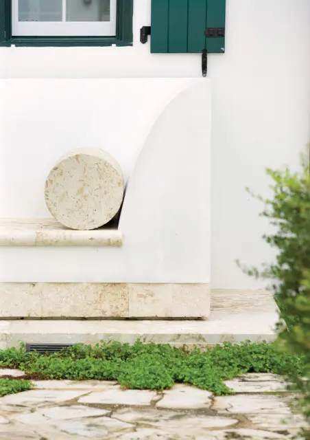 White stucco exterior wall with a curved built-in bench topped with travertine and a cylindrical stone accent. Green shutters flank a window above.