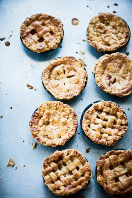Seven golden-brown mini pies with lattice and full crusts arranged on a textured metallic surface.