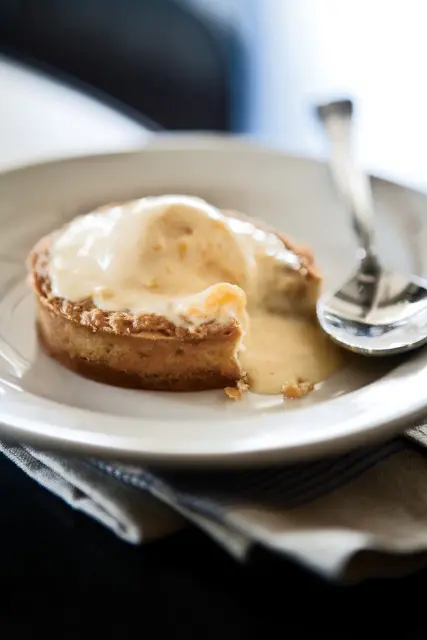 Close-up of a peach tart with vanilla ice cream melting over the top, served on a white plate with a spoon.