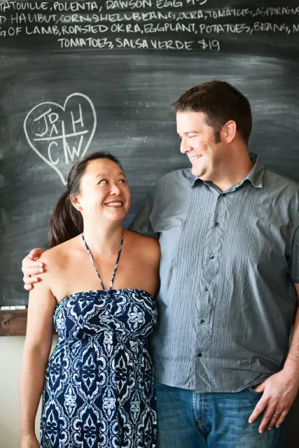 A smiling couple stands in front of a chalkboard menu. The woman wears a blue and white patterned strapless dress, and the man wears a gray button-down shirt and jeans.