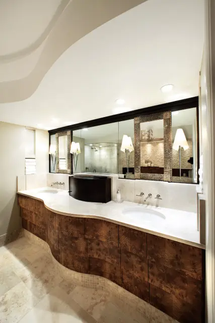 Modern bathroom with a curved vanity featuring a burl wood finish, marble countertop, and dual sinks. A large mirrored wall reflects a walk-in shower and decorative lamps.