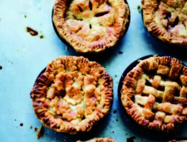 Close-up of four golden-brown mini pies with decorative crusts, two with lattice tops and two with cut-out designs, on a metallic surface.