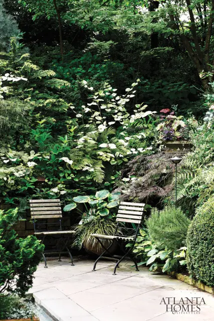 Stone patio with two wooden slatted chairs surrounded by lush green foliage, ferns, and flowering plants.