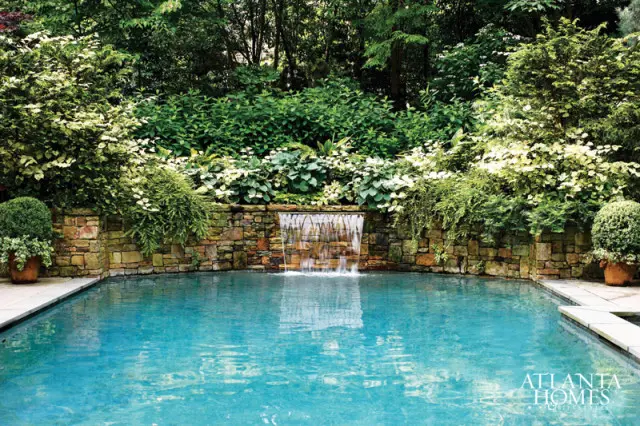 Turquoise swimming pool with a stone waterfall feature surrounded by lush greenery and manicured shrubs.