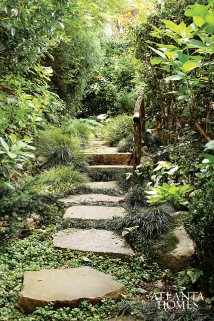 Stone stepping path winding through a lush garden with dense green foliage and ornamental grasses. A rustic wooden railing lines the right side of the path.
