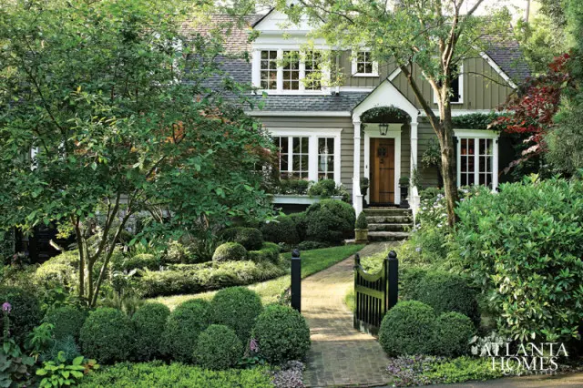 Green clapboard house with white trim, a wooden front door, and a brick pathway leading to the entrance through a manicured garden with boxwood hedges.