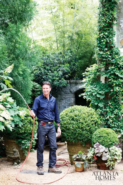 Bob Norris in a blue button-down shirt and jeans holding a hose in a lush garden with bamboo and stone archway in the background.