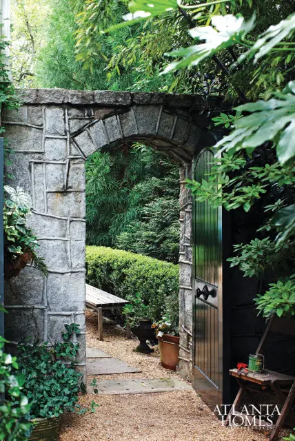 Stone archway leading to a garden path with a wooden bench, manicured hedges, and lush green trees.