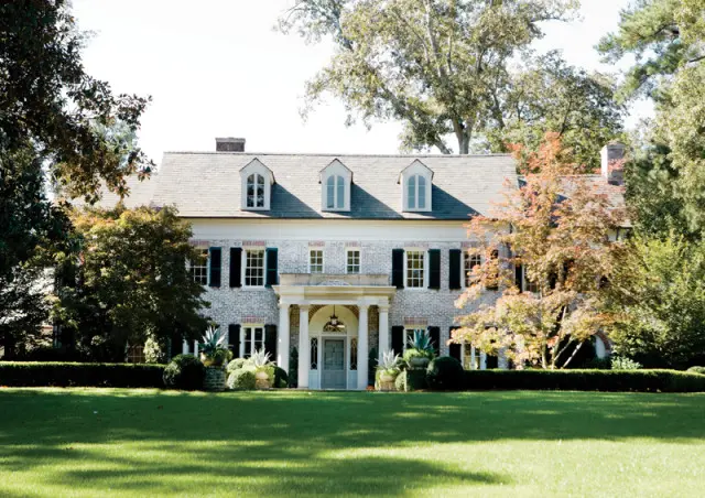 Federal-style white brick house with black shutters, dormer windows, and a columned portico. Lush green lawn and mature trees surround the property.