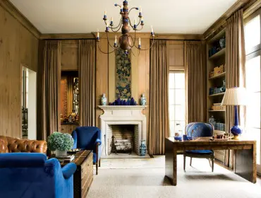Study with wood-paneled walls, a white fireplace, blue velvet armchairs, a leather sofa, a trunk coffee table, and a desk with a blue lamp.