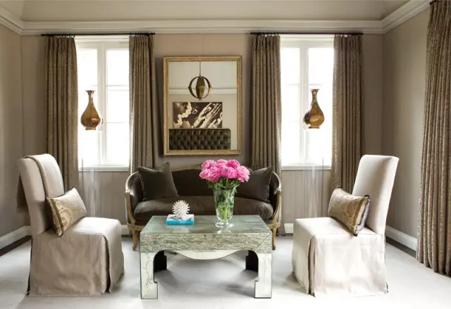 Living room with taupe walls, featuring a dark brown tufted sofa, two cream slip-covered chairs, a mirrored coffee table, and floor-to-ceiling patterned curtains.