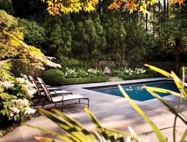 Rectangular swimming pool with blue water surrounded by a stone patio and lush green landscaping, featuring lounge chairs and flowering bushes.