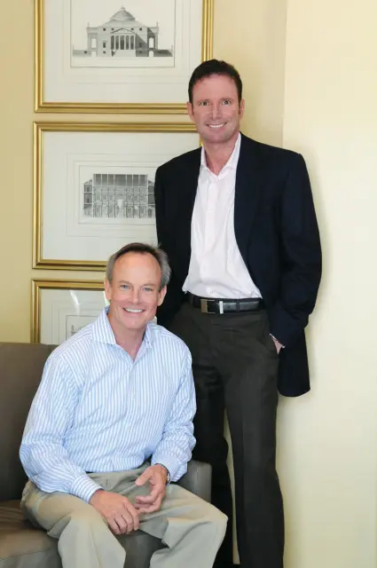 Two men smiling in front of framed architectural drawings. One man sits in a chair wearing a blue and white striped shirt and tan pants. The other man stands wearing a navy blazer, pink shirt, and dark pants.