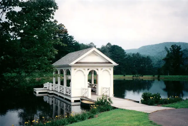 White boathouse with arched openings and decorative railings on a calm lake, surrounded by trees and rolling hills under a cloudy sky.