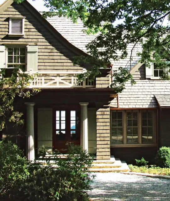 Shingle-sided house with a covered porch, white columns, and a double-door entrance. A decorative white railing adorns the second-story balcony.