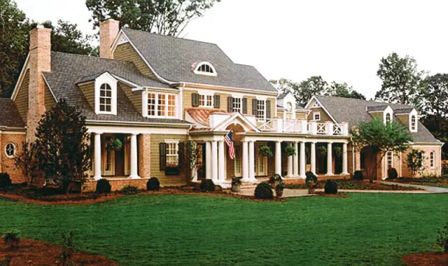 Large Southern Colonial style house with green siding, white columns, brick accents, dormer windows, and a manicured lawn.