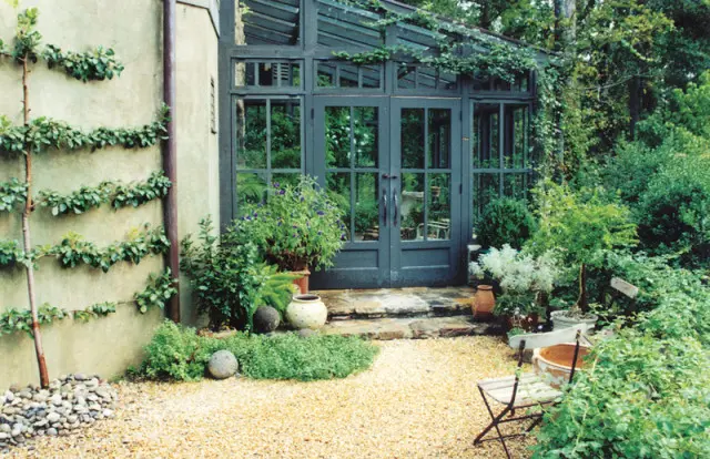 Dark gray greenhouse with double doors and glass panes, surrounded by lush greenery, potted plants, and a gravel pathway. A wall with espaliered plants is visible to the left.