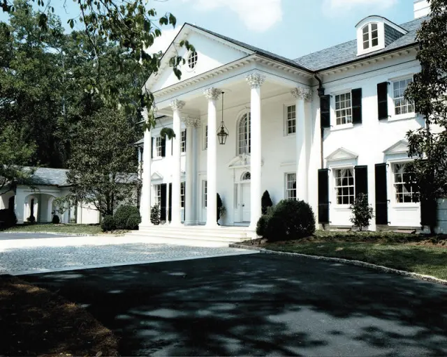 White neoclassical mansion with a grand portico supported by Corinthian columns, black shutters, and a slate roof, surrounded by mature trees and manicured landscaping.