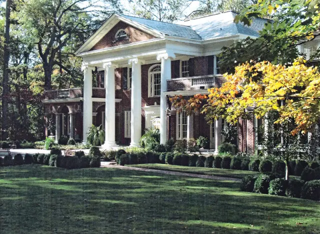 Neoclassical brick house with white columns, arched window, and balcony, surrounded by manicured hedges and a lush green lawn.