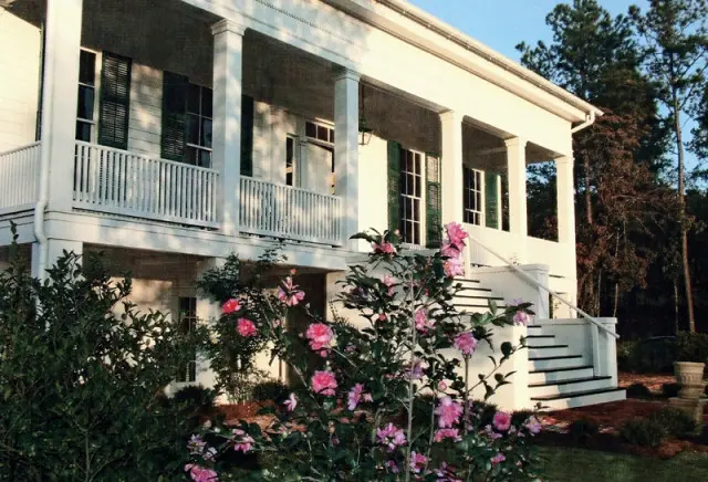 White two-story house with a wraparound porch supported by columns, green shutters, and pink camellia bushes in the foreground.