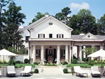 White brick mansion with a covered porch supported by columns, a swimming pool, lounge chairs, and manicured landscaping.