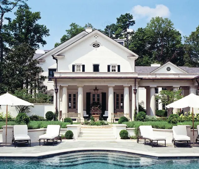 White brick mansion with a covered porch supported by columns, a swimming pool, lounge chairs, and manicured landscaping.