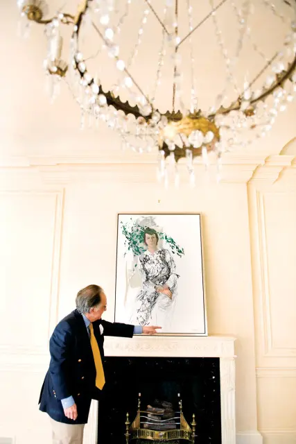 Man in a navy blazer and yellow tie points to a portrait of a woman in a patterned dress hanging above a fireplace. A crystal chandelier hangs from the ceiling.
