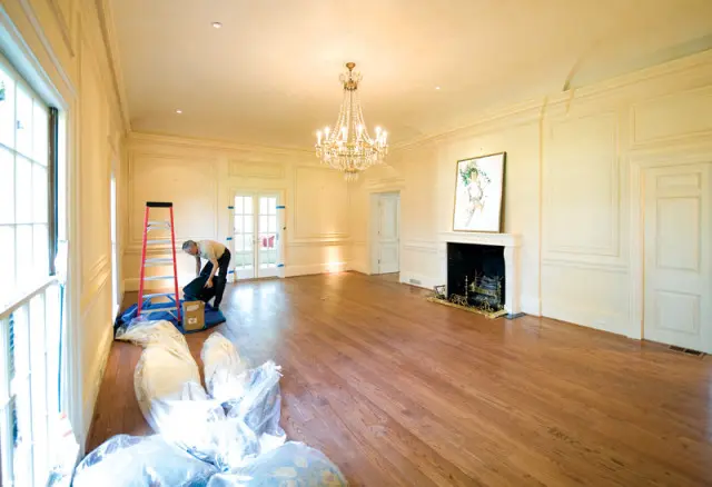 Cream-colored room with hardwood floors, a crystal chandelier, a marble fireplace with a painting above it, and French doors. A man is unpacking items near a ladder.