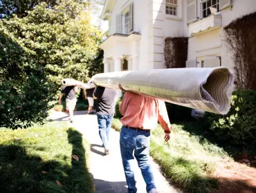 Three men carry a rolled carpet on their shoulders along a stone path next to a white house with green landscaping.