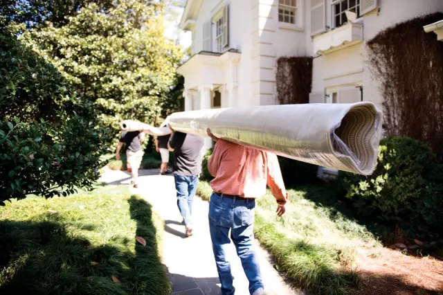 Three men carry a rolled carpet on their shoulders along a stone path next to a white house with green landscaping.